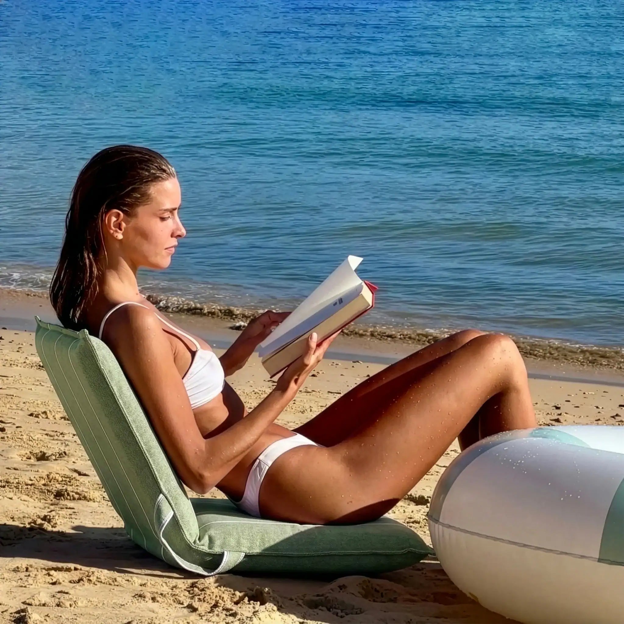 Woman reading on a green La Chaise portable reclining beach lounger by the water